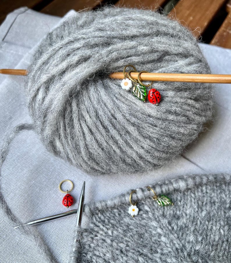 K&ouml;nnte beinhalten: Graue Garnkugel mit einer Stricknadel aus Holz und drei Maschenmarkern. Die Maschenmarker sind ein Marienk&auml;fer, eine Blume und ein Blatt. Das Garn liegt auf einer grauen Oberfl&auml;che.