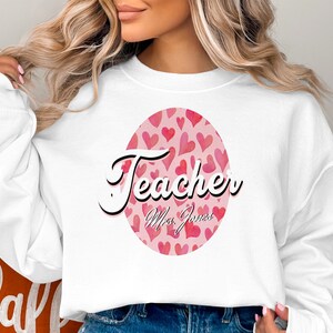 May include: White sweatshirt with a pink circle design featuring a pattern of pink hearts and the text "Teacher" with the name "Mrs. Jones" underneath.