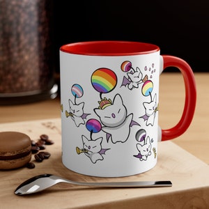 May include: White ceramic mug with a red rim and handle. The mug features a colourful design of cartoon bats with rainbow and pride flags.