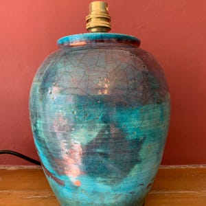 May include: A ceramic table lamp with a turquoise and blue glaze. The lamp has a crackle finish and a brass lamp holder. The lamp is on a wooden surface.