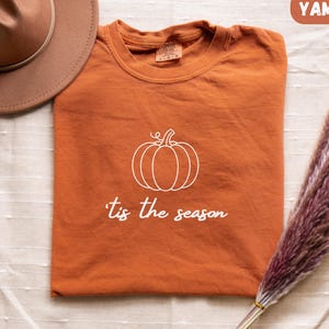 Peut inclure: Un t-shirt à col rond orange brûlé avec un motif de citrouille en ligne blanche et le texte 'tis the season imprimé en blanc. Le t-shirt est présenté avec un chapeau marron à larges bords et un bouquet de tiges d'herbe de la pampa séchées.