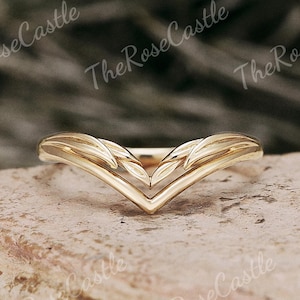 Puede incluir: Un anillo dorado con un diseño en forma de V. El anillo presenta patrones en forma de hojas en la parte superior, creando un aspecto delicado. El anillo se encuentra sobre un fondo neutro, resaltando sus intrincados detalles. Una joya.