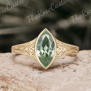 May include: A gold ring with a marquise-cut, light green gemstone. The band features an ornate, engraved design. The ring is set on a light brown surface with a blurred background.
