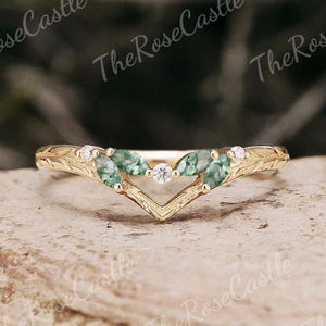 Può includere: Un anello color oro con un design a forma di V. L'anello presenta tre pietre preziose verdi e piccole gemme bianche. La fascia ha un motivo strutturato e inciso. L'anello è posto su uno sfondo sfocato.