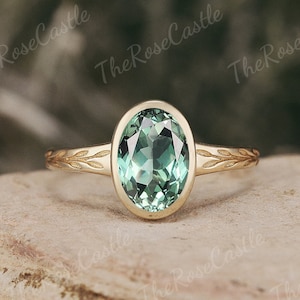 May include: A gold ring featuring an oval, pale green gemstone. The ring's band is adorned with a leaf pattern. The gemstone is set in a bezel setting. The ring is displayed on a stone surface.