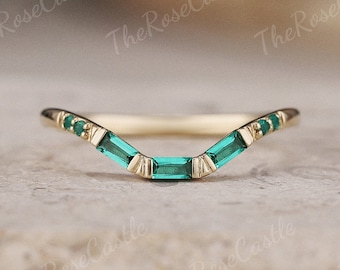 May include: A curved gold ring with emerald-cut green gemstones. The ring has a delicate design with a series of emerald-cut stones set in a curved pattern. The ring is set against a neutral background.