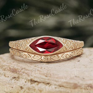 May include: A gold-coloured ring set with a marquise-cut, deep red gemstone. The rings feature intricate floral and scrollwork designs. The text "The Rose Castle" is visible in the background.