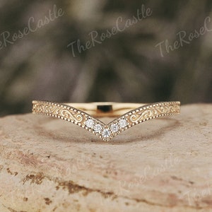 Może przedstawiać: Delikatny złoty pierścionek z wzorem w kształcie litery V i małymi diamentami osadzonymi w środku. Obrączka jest ozdobiona misternymi zdobieniami.