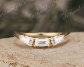 May include: A gold ring with three white baguette-cut stones. The center stone is rectangular, flanked by two trapezoid-shaped stones. The ring is set on a light brown surface. The Rose Castle is printed on the background.