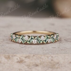 Puede incluir: Un delicado anillo de banda de oro con una fila de piedras preciosas verdes y blancas alternadas. Las piedras preciosas verdes son de forma baguette y las piedras preciosas blancas son redondas.