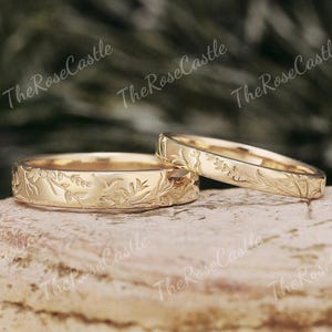 May include: Two gold wedding bands with floral engravings. The rings are displayed on a light brown surface. The text "The Rose Castle" is visible in the background.