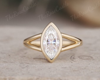 Puede incluir: Un anillo de oro con un diamante talla marquesa. El diamante está engastado en un bisel y la banda se divide en dos líneas paralelas a cada lado del diamante. El anillo se muestra sobre una superficie neutra.