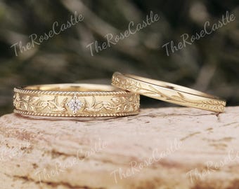 May include: Two gold wedding rings. One ring features a central diamond and intricate floral and leaf detailing. The other ring has a delicate leaf pattern. The rings are displayed on a textured surface.