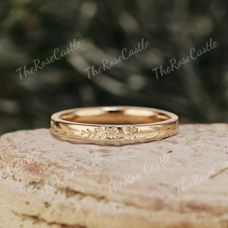 K&ouml;nnte beinhalten: Ein goldfarbener Ring mit floralem Design. Der Ring hat ein eingraviertes Muster aus Blumen und Bl&auml;ttern um das Band. Er wird auf einer strukturierten, hellbraunen Oberfl&auml;che pr&auml;sentiert. Der Text "The Rose Castle" ist im Hintergrund sichtbar.
