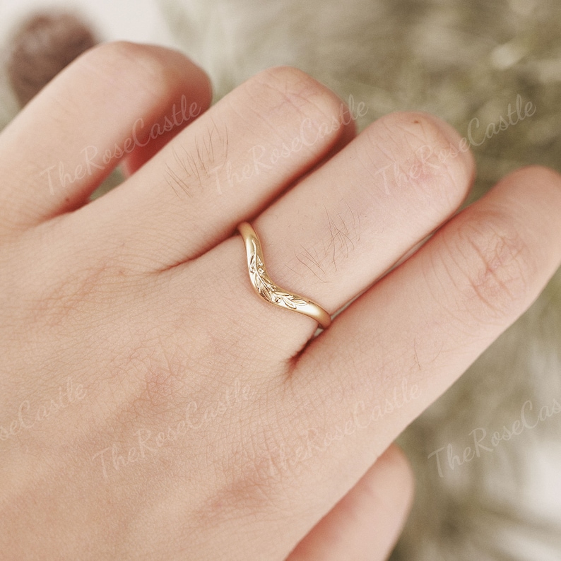 May include: A gold-colored ring with a V-shaped design and floral details. The ring is worn on a finger.