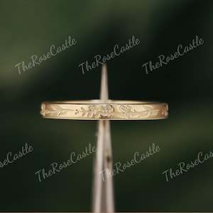 K&ouml;nnte beinhalten: Ein goldfarbener Ring mit floralem Muster. Der Ring hat ein sich wiederholendes Design aus Blumen und Bl&auml;ttern um das Band. Der Ring wird vor einem verschwommenen gr&uuml;nen Hintergrund pr&auml;sentiert. Der Text "The Rose Castle" ist im Hintergrund sichtbar.