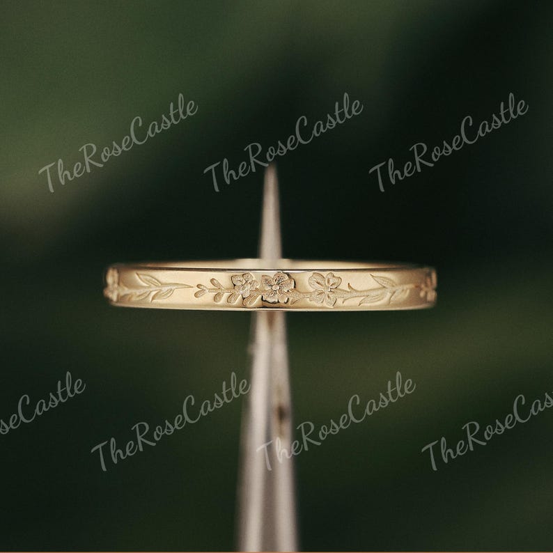 K&ouml;nnte beinhalten: Ein goldfarbener Ring mit floralem Muster. Der Ring hat ein sich wiederholendes Design aus Blumen und Bl&auml;ttern um das Band. Der Ring wird vor einem verschwommenen gr&uuml;nen Hintergrund pr&auml;sentiert. Der Text "The Rose Castle" ist im Hintergrund sichtbar.