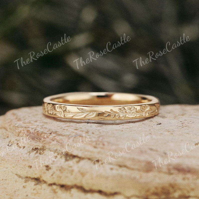 K&ouml;nnte beinhalten: Ein goldfarbener Ring mit einem floralen und Blattmuster, das um das Band graviert ist. Der Ring liegt auf einer Steinoberfl&auml;che. Der Text "The Rose Castle" ist im Hintergrund wiederholt.