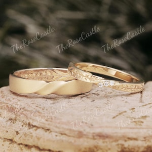 May include: Two gold wedding rings. One ring has a twisted design with a matte finish and engraved floral pattern. The other ring has a floral pattern with three small diamonds. The rings are displayed on a stone surface.