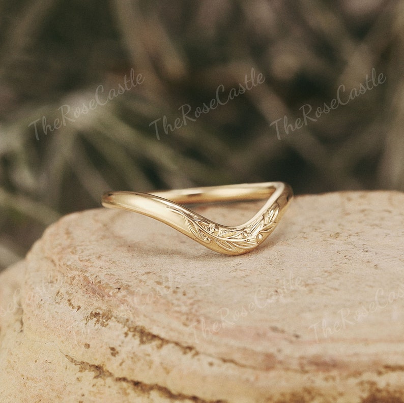 May include: A delicate gold ring with a curved, v-shaped band and a floral design. The ring is set on a textured, light brown surface.