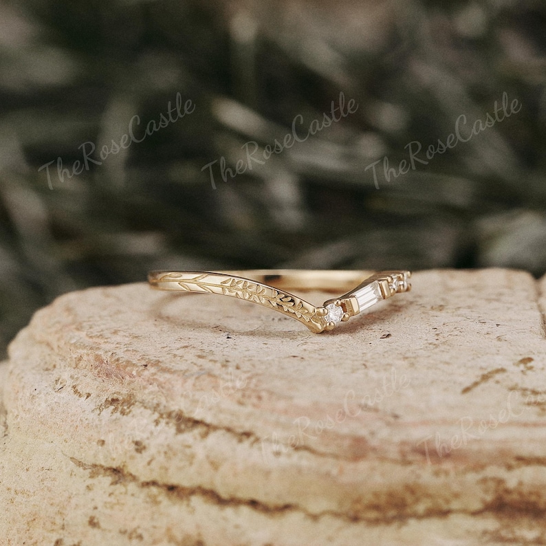 May include: A delicate gold band with a v-shaped design and a small diamond set in the center. The band is adorned with a floral pattern.
