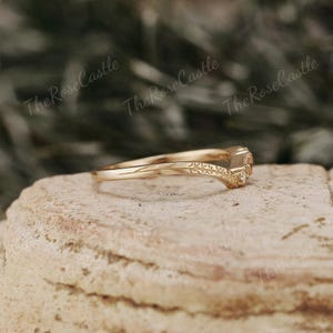 May include: A gold band ring with a delicate, curved design and three small, clear gemstones. The ring is set on a textured, brown surface.