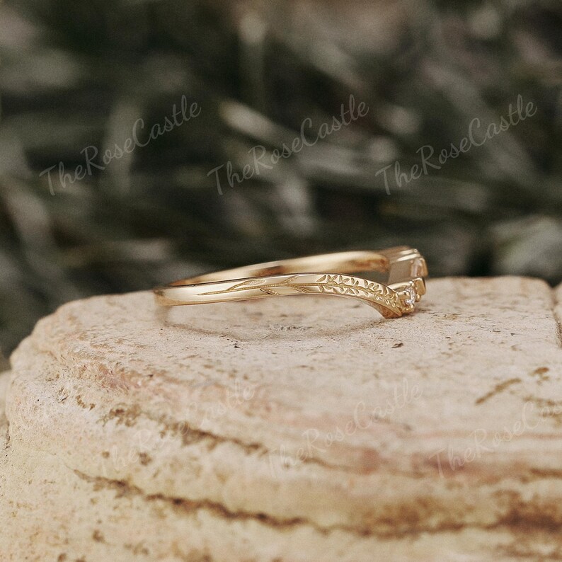 May include: A gold band ring with a delicate, curved design and three small, clear gemstones. The ring is set on a textured, brown surface.