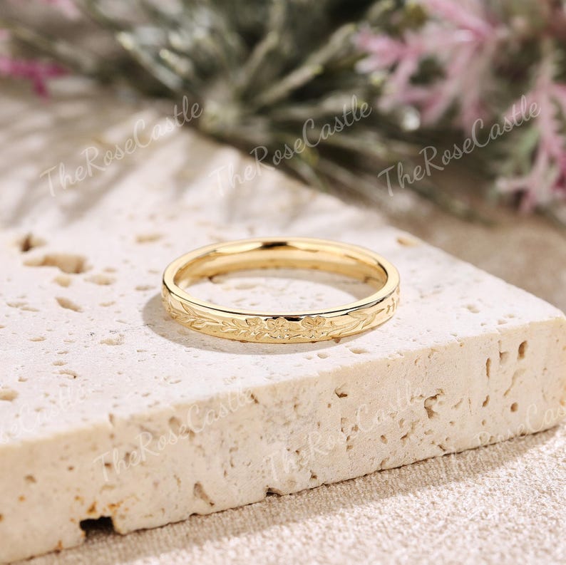 K&ouml;nnte beinhalten: Ein goldfarbener Ring mit floralem Muster. Der Ring liegt auf einer hellen, strukturierten Oberfl&auml;che. Das Design des Rings zeigt zarte Bl&auml;tter und Bl&uuml;ten, die eine elegante und zeitlose &Auml;sthetik schaffen. Der Ring ist ein Symbol f&uuml;r Engagement und Liebe.
