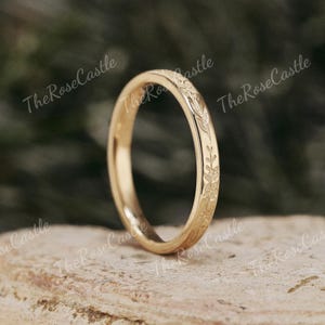 K&ouml;nnte beinhalten: Ein goldfarbener Ring mit einem floralen Muster, das um das Band graviert ist. Der Ring ist vor einem neutralen Hintergrund platziert. The Rose Castle ist auf dem Bild aufgedruckt.