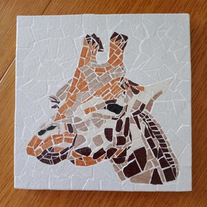 Op de afbeelding: Een vierkante mozaïektegel met de kop van een giraffe. De giraffe is weergegeven in tinten bruin, beige en wit, tegen een witte achtergrond. Het mozaïek is samengesteld uit kleine, onregelmatig gevormde tegels, wat een textuureffect geeft.