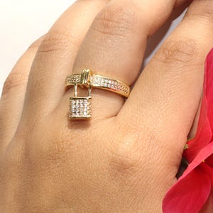Könnte beinhalten: Ein goldfarbener Ring mit einem Anhänger in Form eines Vorhängeschlosses. Das Ringband ist mit kleinen, klaren Steinen besetzt. Der Vorhängeschloss-Anhänger ist quadratisch und ebenfalls mit kleinen, klaren Steinen bedeckt.
