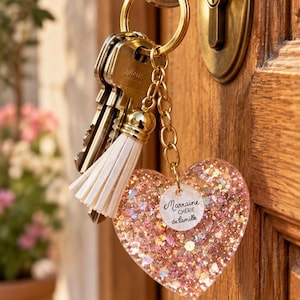 Op de afbeelding: Een goudkleurige sleutelhanger met een hartvormige bedel gevuld met roze en gouden glitters, een witte kwast en een klein rond label met de tekst Marraine CHÉRIE de Camille, bevestigd aan een sleutel in een houten deurslot.