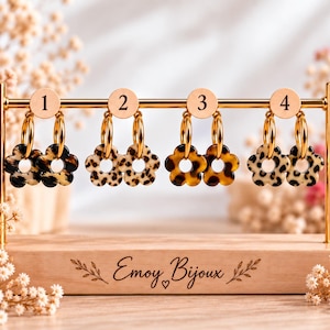 Op de afbeelding: Vier paar gouden oorringen met bloemvormige bedels in diverse luipaardprints, genummerd van één tot vier op een gouden displayrek. De houten voet is gegraveerd met de tekst Emoy Bijoux en decoratieve bladaccenten.