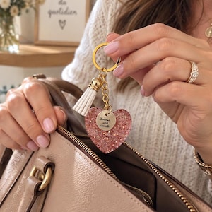 Op de afbeelding: Een goudkleurige sleutelhanger met een hartvormige bedel gevuld met roze glitters en een kleine ronde gouden schijf met de inscriptie "Immer und überall". De sleutelhanger bevat een witte kunstlederen kwast en een gouden metalen ring.