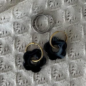 Pode incluir: Dois pares de brincos, um par é de argolas de prata e o outro par é de argolas de ouro com pingentes de flores pretas.