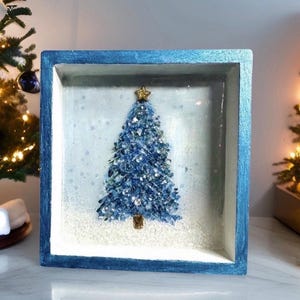 May include: A square shadow box frame with a blue painted border containing a textured Christmas tree made of blue and silver crystalline pieces. The tree is topped with a small gold star and sits on a white base resembling snow.