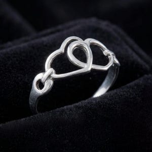 May include: A silver ring featuring a linked heart design. The ring has three connected hearts, with a polished finish. The ring is displayed on a black velvet surface.
