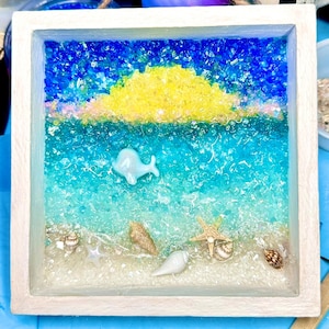 May include: A white wooden frame with a glass front filled with blue, yellow, and white glass beads. The beads are arranged to look like a beach scene with a blue sky, yellow sun, and blue water. There are also small white seashells and a starfish in the scene.