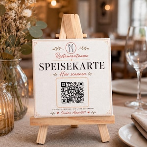 Könnte beinhalten: Ein quadratisches weißes Schild mit schwarzem Text und einem QR-Code auf einer kleinen Holzstaffelei. Das Schild enthält den Text Restaurantname, SPEISEKARTE, Hier scannen, FRISCH. REGIONAL. MIT LIEBE ZUBEREITET. und Guten Appetit! mit Symbolen.