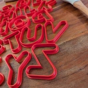 Könnte beinhalten: Ein Satz roter Kunststoff-Ausstecher in Form von Buchstaben des Alphabets. Die Ausstecher sind auf einer Holzoberfläche gestapelt.