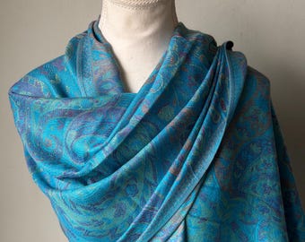 May include: A draped, turquoise paisley scarf on a white mannequin head. The scarf features intricate patterns in shades of blue, purple, and teal. The fabric appears soft and flowing, with a subtle sheen. The scarf is a fashion accessory.