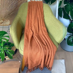 Op de afbeelding: Een oranje-bruine deken met franjes op een groene fauteuil. De deken is gevouwen en over de stoel gelegd.