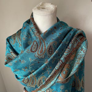 May include: A teal paisley scarf draped over a white mannequin head. The scarf features a detailed pattern in shades of brown and gold. The edges are trimmed with a darker brown border, adding a touch of elegance to the textile.