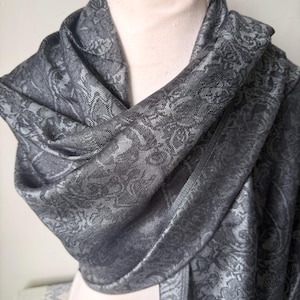 Könnte beinhalten: Dunkelgrauer Schal mit Paisley-Muster, drapiert auf einer Schaufensterpuppe. Der Schal hat ein detailliertes Blumendesign und besteht aus einem weichen, fließenden Stoff. Der Schal ist um den Hals und die Schultern der Schaufensterpuppe gewickelt.