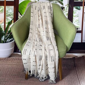May include: A cream-colored throw blanket with a black geometric pattern draped over a green armchair. The blanket has a fringe border.