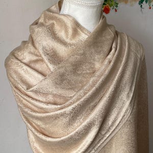 Puede incluir: Un chal beige con estampado de cachemira drapeado sobre un maniquí. El chal está hecho de una tela suave y fluida y presenta un diseño detallado en toda la superficie. Los bordes están acabados con un borde sutil.