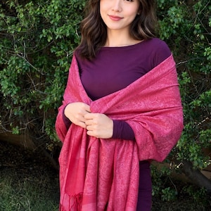 Op de afbeelding: Een magenta paisley sjaal met een gefranjerde rand. De sjaal is gedrapeerd over een donkerpaarse jurk met lange mouwen. Het ingewikkelde patroon en de levendige kleur van de sjaal creëren een opvallend contrast met de jurk. De afbeelding is tegen een achtergrond van groen gebladerte.