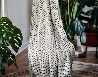 May include: A white throw blanket with a black geometric pattern of dots and arrows. The blanket has a fringe border and is draped over a chair.
