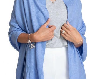 May include: A light blue shawl draped over a gray top and white pants. The shawl has a textured appearance. The woman is wearing a necklace and bracelets.