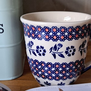 May include: A white ceramic mug with a blue and pink floral pattern. The mug has a blue handle and a wide, rounded base.